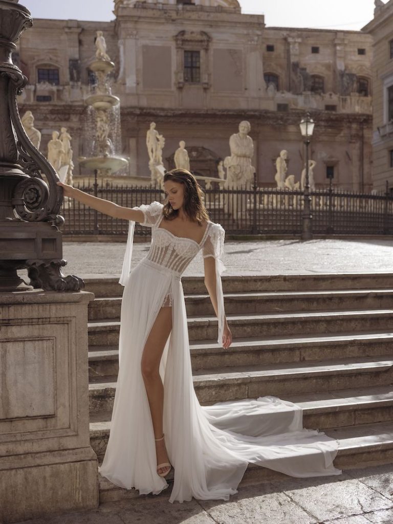 In questa foto una modella posa davanti a piazza Pretoria, a Palermo, indossando un vestito con profondo spacco e dolci maniche corte a palloncino. L'abito è firmato Pinella Passaro 2023