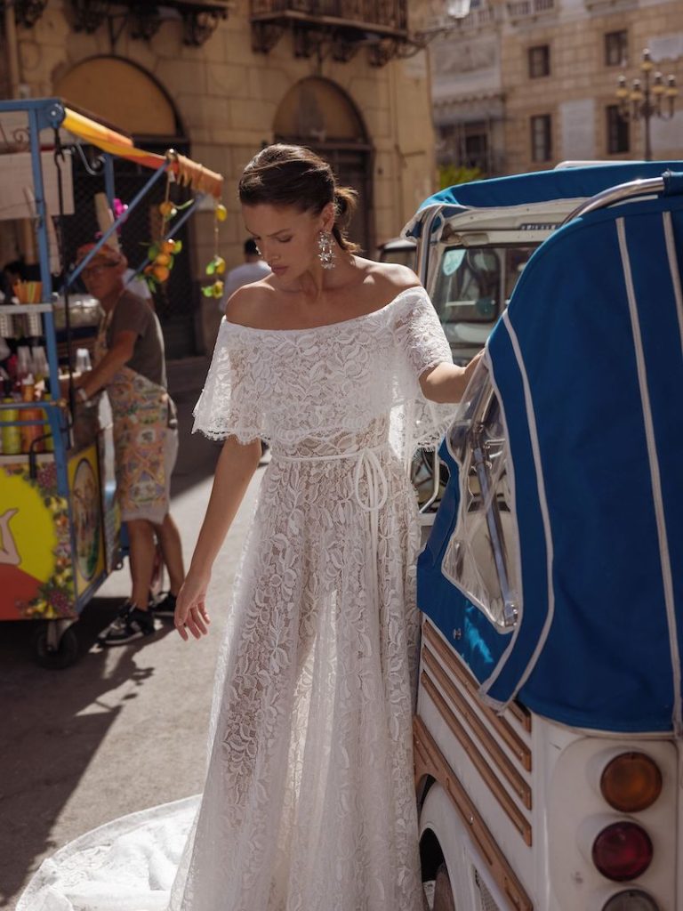 In questa foto una modella indossa un leggerissimo abito da sposa in pizzo, con scollatura omerale, della collezione Pinella Passaro 2023