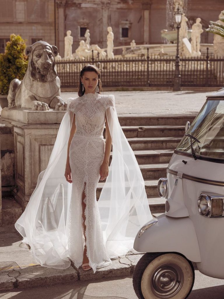 In questa foto una modella indossa un abito da sposa in pizzo all over. Particolari le ali di tessuto che si diramano dalle spalline rettangolari. Il vestito fa parte della collezione sposa Pinella Passaro 2023