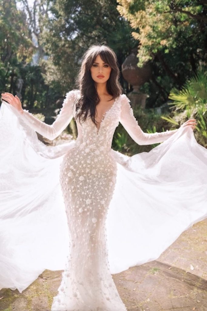 In questa foto una modella indossa un abito da sposa Dovita Bridal 2023 a sirena color champagne con scollo a V, maniche lunghe e con ricami di fiori 3D e perline. La modella tiene tra le mani la sovragonna di tulle