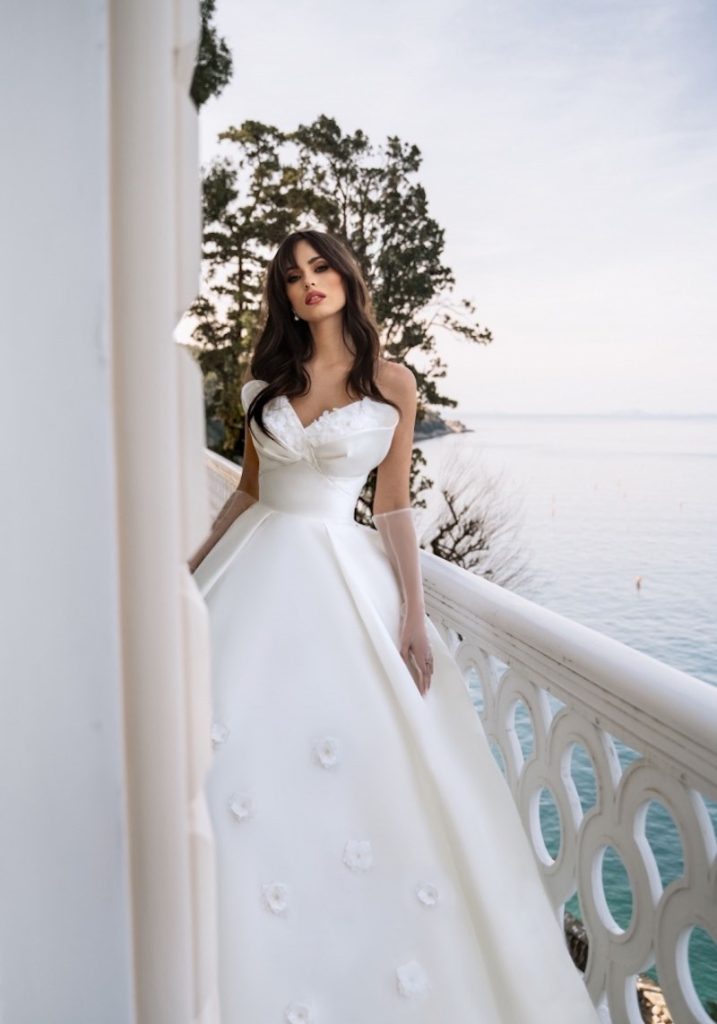 In questa foto una modella indossa un abito da sposa Dovita Bridal 2023 ampio in chiffon con fiori 3D sulle lunghezze, cintura stretta in vita e voluminosa scollatura a fiore. La modella indossa anche guanti in tulle trasparente