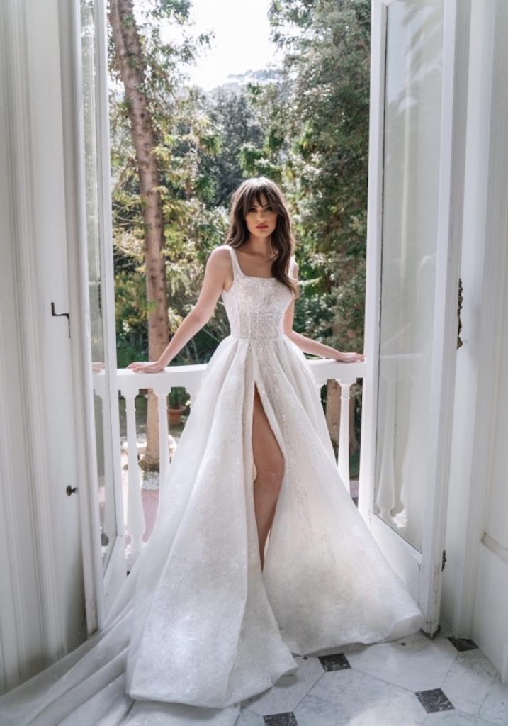 In questa foto una modella indossa un abito da sposa Dovita Bridal 2023 in pizzo ricamato ampio con spacco frontale e scollo quadrato