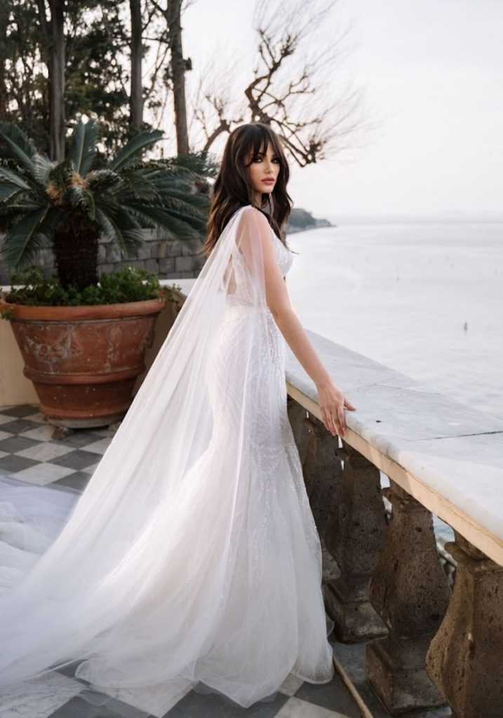 In questa foto una modella indossa un abito da sposa Dovita Bridal 2023 a sirena in pizzo ricamato con paillettes. Dalle spalle scendono due ali di tulle rimovibili