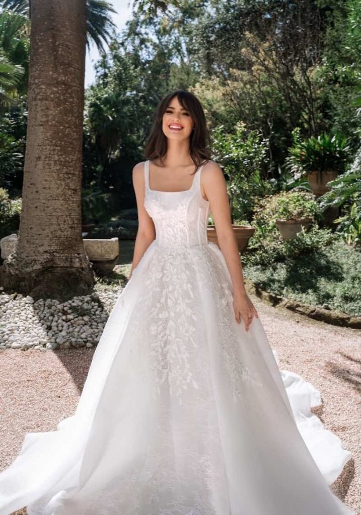 In questa foto una modella indossa un abito da sposa Dovita Bridal 2023 dalla linea ad A in pizzo Chantilly con scollatura quadrata e ramage floreale sul corpetto strutturato e sulle lunghezze