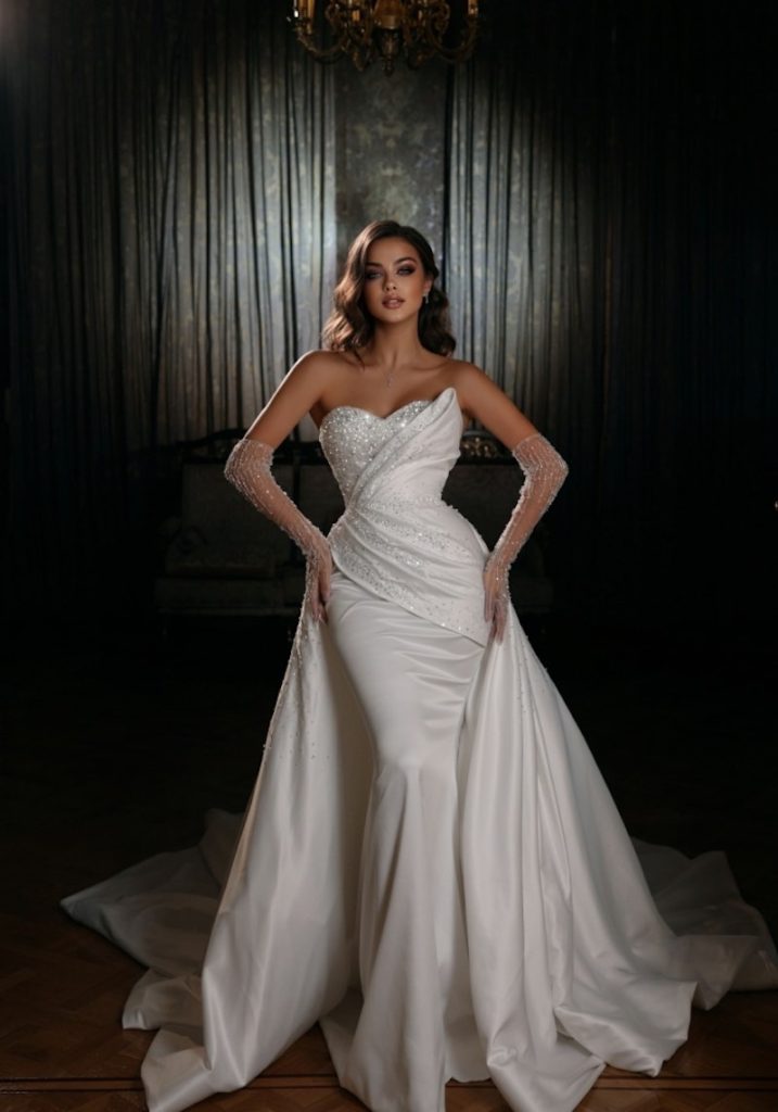 In questa foto una modella indossa un abito da sposa Dovita Bridal 2023 a sirena con sopragonna in raso di seta. Il corpetto è ricamato a metà con cristalli. La modella indossa anche guanti di tulle trasparente con cristalli