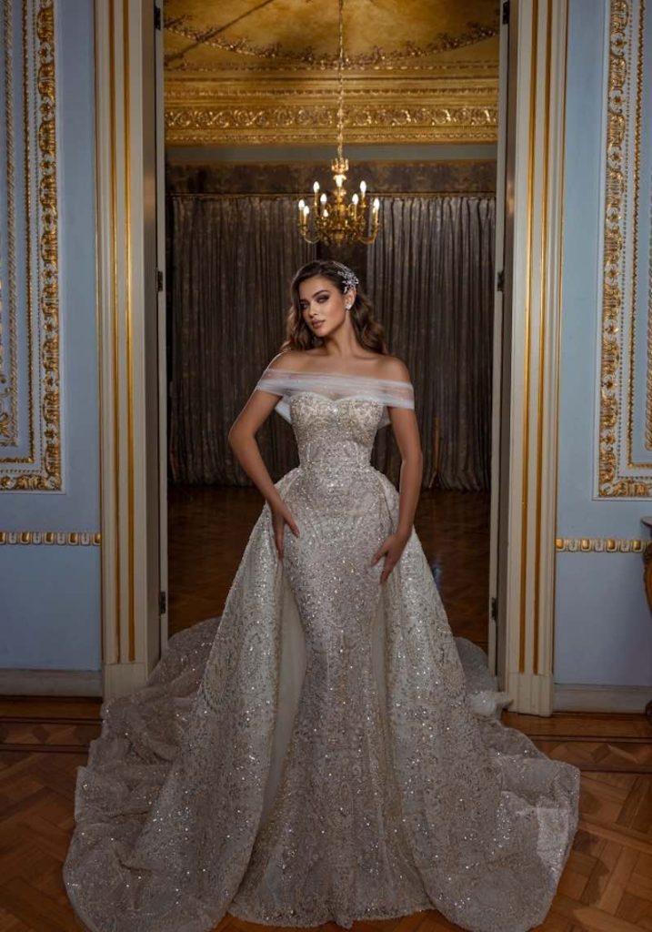 In questa foto una modella indossa un abito da sposa Dovita Bridal 2023 a sirena con sopragonna staccabile interamente ricamati di paillettes e perline. La sposa indossa anche una stola di tulle trasparente