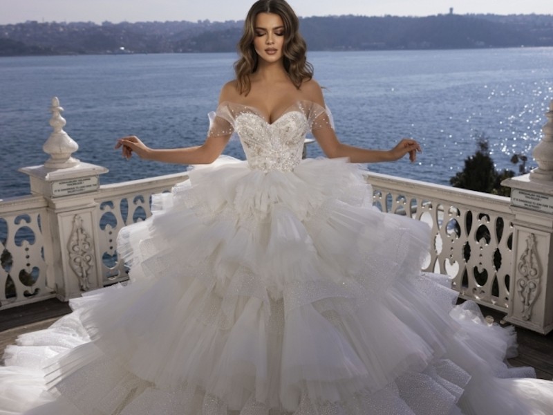 In questa foto una modella indossa un abito da sposa Dovita Bridal 2023 Premium voluminoso con maxi ruches di tulle su tutta la lunghezza e corpetto a cuore ricamato con cristalli. Sulle spalle sono prese fasce di tulle off-shoulder con applicazioni di cristalli