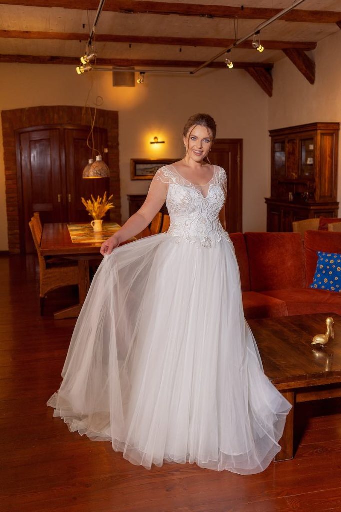 In questa foto una modella indossa un abito da sposa curvy scivolato con gonna in tulle e corpetto a maniche corte con ricamati in rilievo