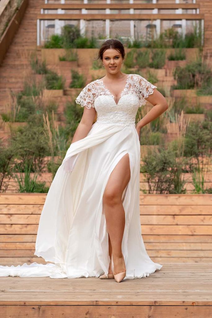 In questa foto una modella indossa un abito da sposa curvy scivolato con scollo a V e spacco in chiffon con corpetto a maniche corte in pizzo 