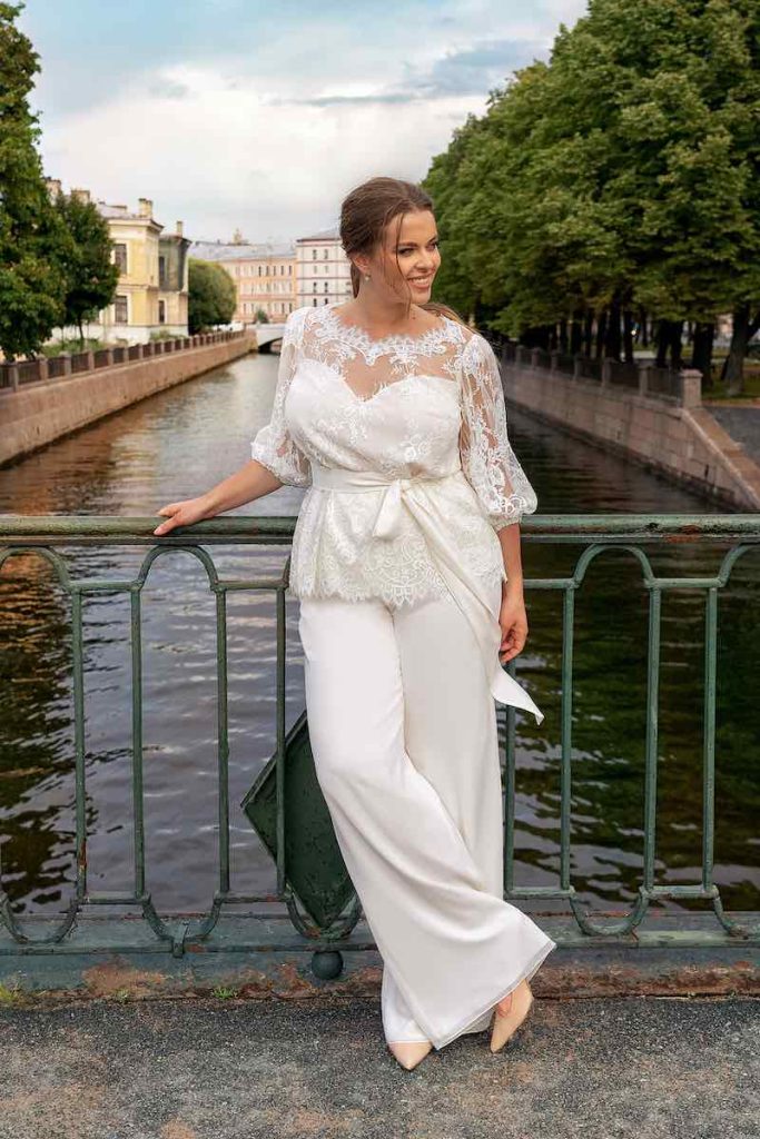 In questa foto una modella posa in riva al fiume indossando un abito da sposa con pantaloni e blusa in pizzo a maniche a sbuffo a tre quarti in pizzo