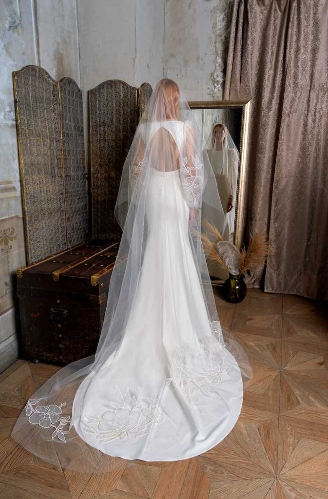 In questa foto una modella posa di spalle davanti ad uno specchio indossando un abito da sposa Papilio 2023 a mezza sirena in georgette con scollatura sulle schiena e velo medio in tulle ricamato a taglio nudo