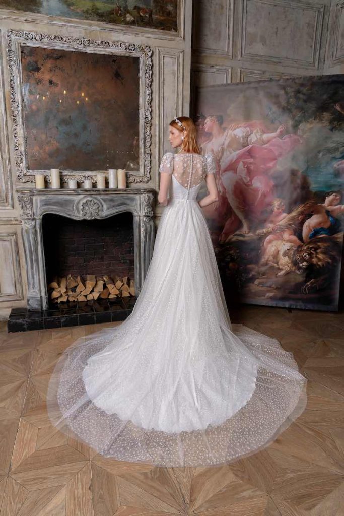 In questa foto una modella ritratta di spalle indossa un abito da sposa scivolato Papilio 2023 in georgette e lungo strascico in tulle plumetis. L'abito presenta  maniche corte a palloncino chiusura posteriore con bottoni