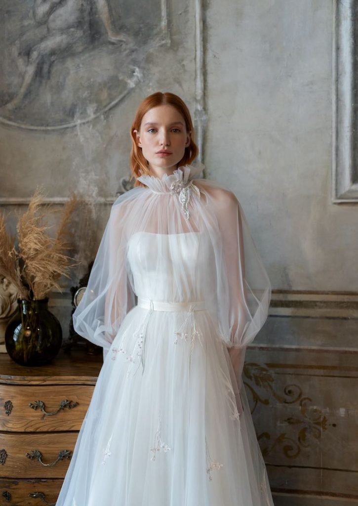 In questa foto una modella indossa un abito da sposa scivolato in tulle con cappa arricciata al collo con maniche lunghe a sbuffo