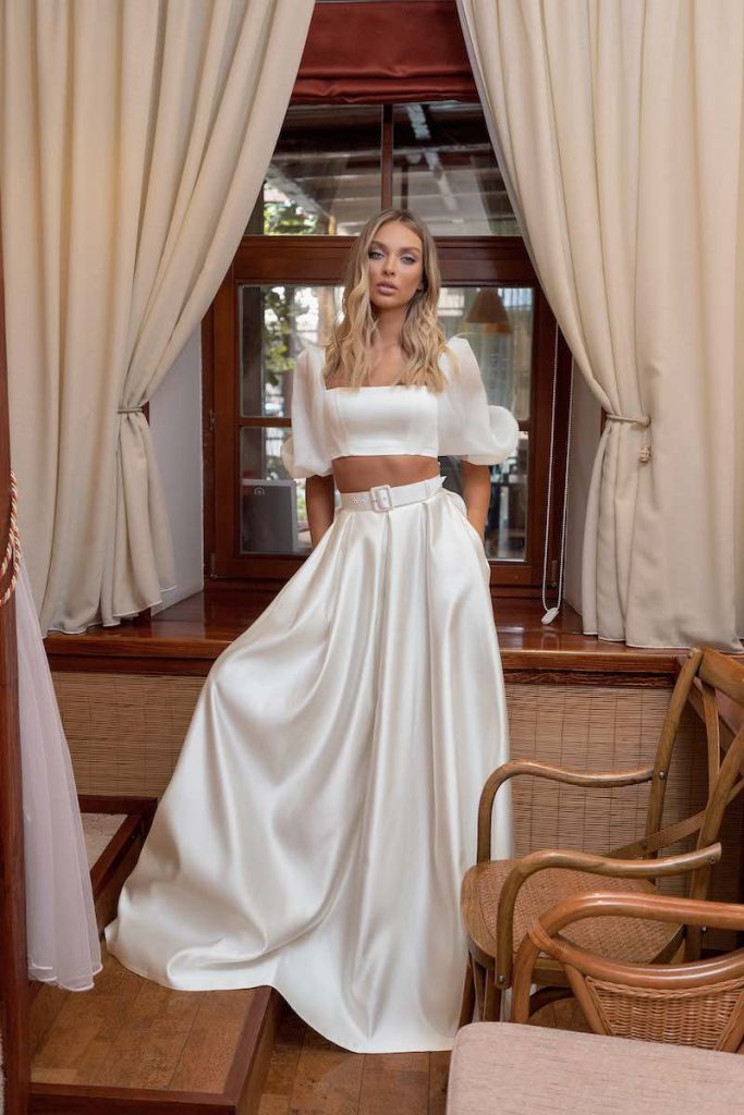 In questa foto una modella indossa un abito da sposa componibile Papilio 2023 con crop top con maniche a tre quarti a sbuffo e gonna in raso di seta con cinturone 