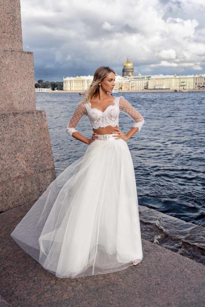 In questa foto una modella posa in riva al mare indossando un abito da sposa Papilio 2023 composto da un crop top con maniche a tre quarti in tulle plumetis e pizzo e una gonna in tulle fermata da un cinturone