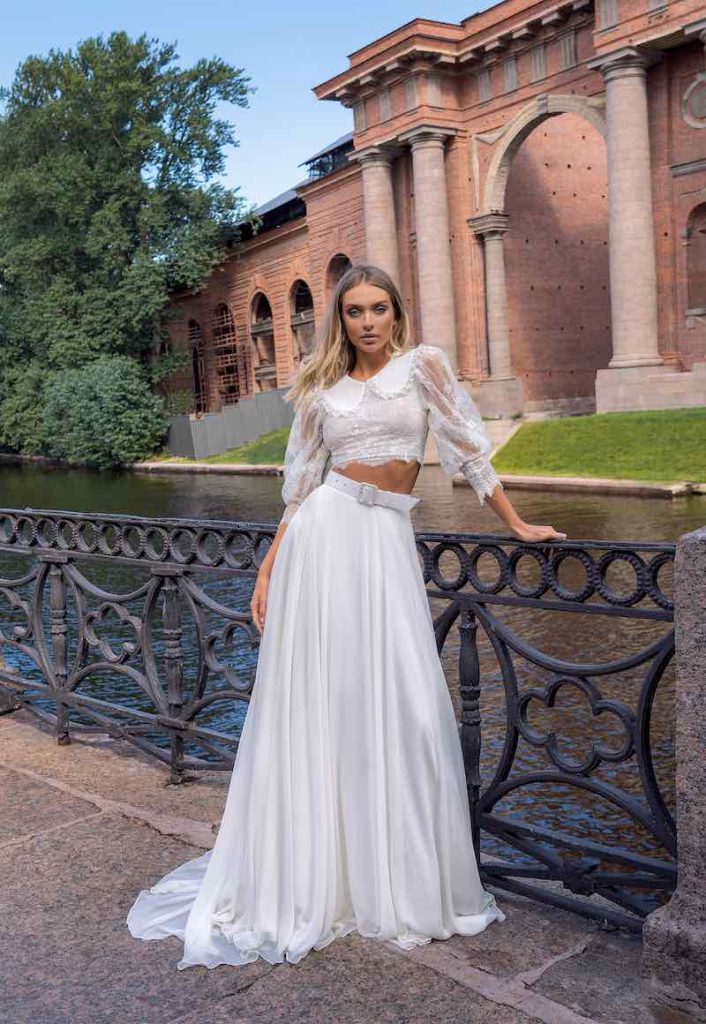 In questa foto una modella indossa un abito da sposa componibile Papilio 2023 con crop to di pizzo con maniche lunghe e colletto e gonna lunga in chiffon con cinturone