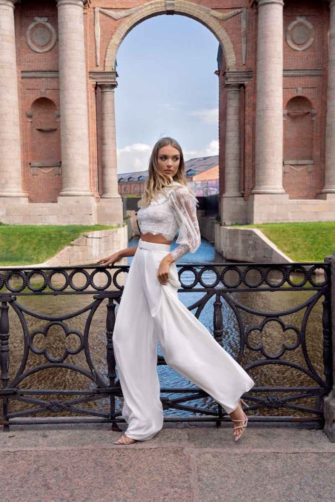 In questa foto una modella indossa un abito da sposa con crop top in pizzo e maniche lunghe a sbuffo e pantaloni ampi in georgette