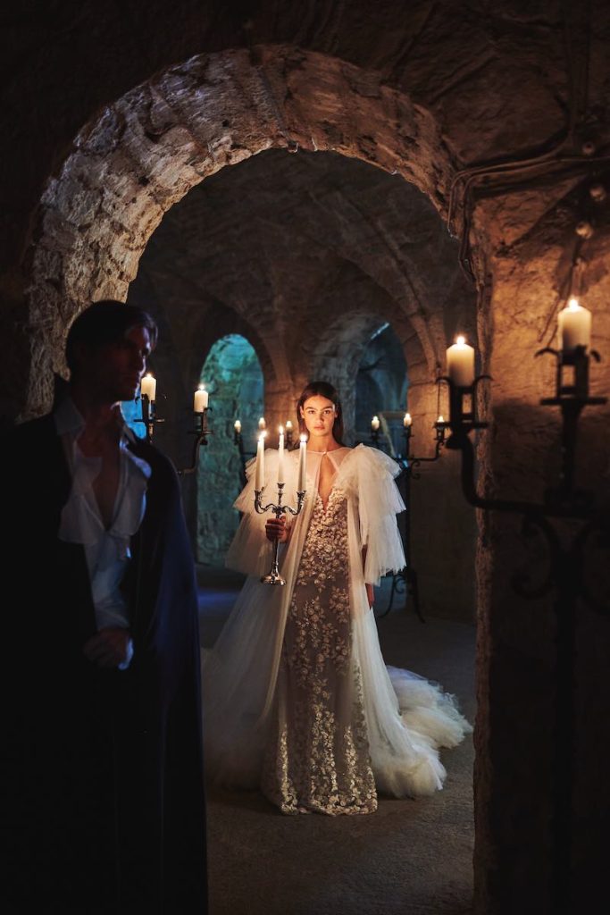 In questa foto una modella posa al buio in un castello indossando un abito da sposa a sirena Alessandro Angelozzi Couture 2023 in pizzo floreale abbinato ad una mantella in tulle a balze legata al collo