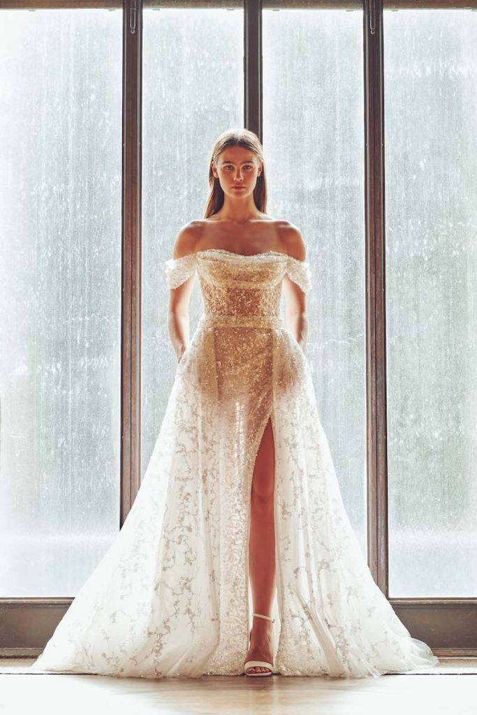 In questa foto una modella indossa un abito da sposa in tulle sparkling Alessandro Angelozzi Couture 2023 con spacco frontale e maniche off shoulder