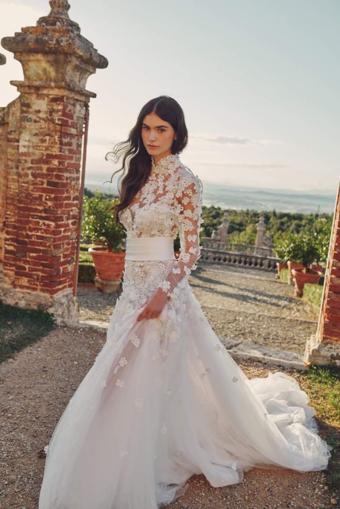 In questa foto una modella indossa un abito da sposa Alessandro Angelozzi Couture 2023 scivolato in tulle con maniche e applicazioni di pizzo floreale 3D sul corpetto e sulle lunghezze. L'abito in vita presenta anche una cintura in mikado leggermente drappeggiato