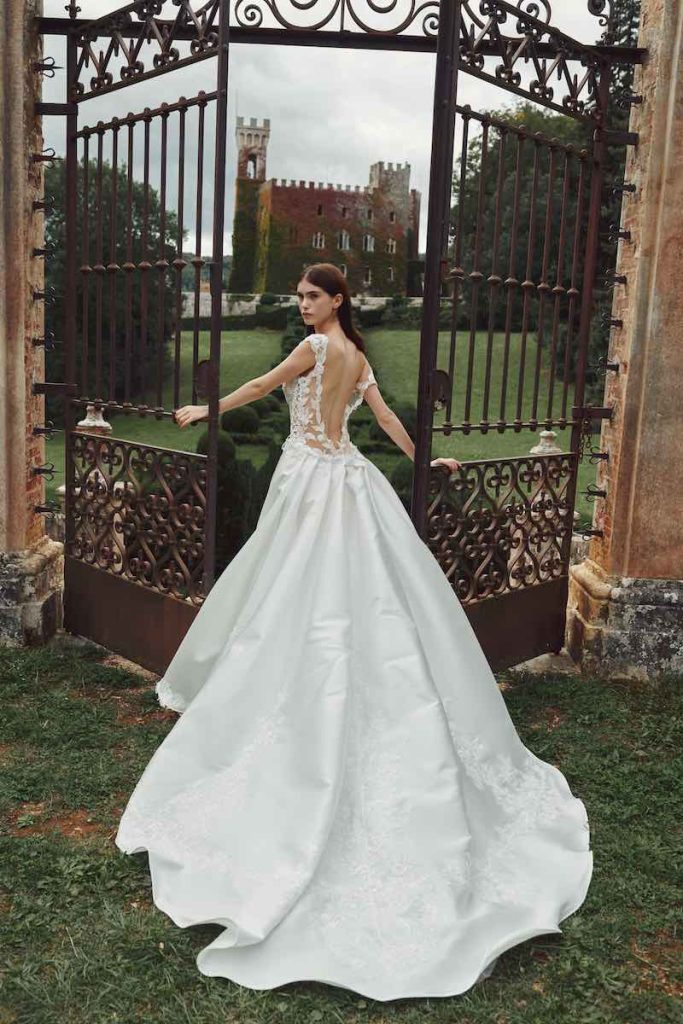 In questa foto una modella posa uscendo da un cancello in ferro davanti ad un castello indossando un abito da sposa Redingote Alessandro Angelozzi Couture 2023 con una lunga coda in mikado e schiena scoperta incorniciata da pizzo chantilly 