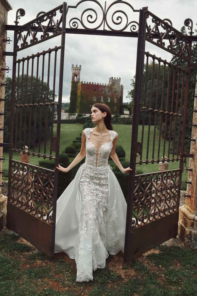 In questa foto una modella posa uscendo da un cancello davanti a un castello con un abito da sposa Alessandro Angelozzi Couture 2023 in pizzo, scollo a cuore e sopragonna in chiffon. L'abito presenta anche maniche corte in pizzo