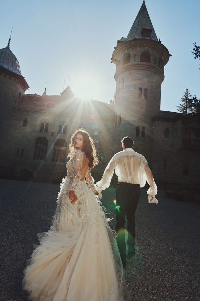 In questa foto una modella posa di spalle correndo per mano con un modello e indossando un abito da sposa Alessandro Angelozzi Couture 2023 in tulle e piume con schiena scoperta