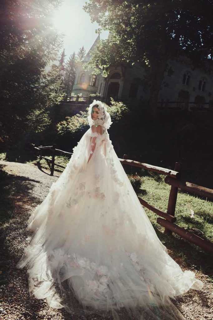 In questa foto una modella posa correndo indossando una mantella da sposa in tulle con cappuccio e applicazione di fiori all over 