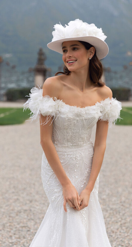 In questa foto una modella indossa un abito da sposa Pollardi 2023. Il dettaglio di stile è il cappello con applicazione di fiori in tessuto dalle dimensioni maxi