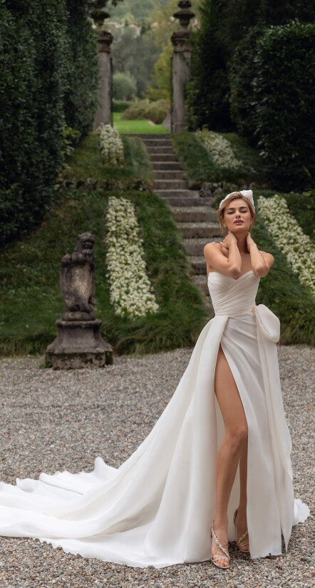 In questa foto una modella, in un giardino, indossa un abito con corpetto con scollo a cuore e un profondissimo spacco. Il vestito fa parte della collezione sposa Pollardi 2023