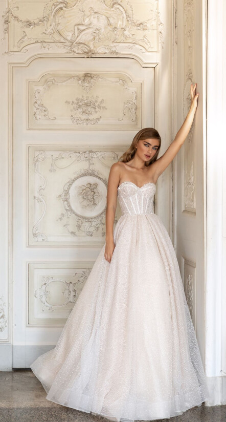 In questa foto una modella indossa un abito da sposa romantico, con corpetto steccato con scollatura a cuore e gonna vaporosa e scivolata. Il tessuto ha un leggero effetto glitterato, con un colore rosato