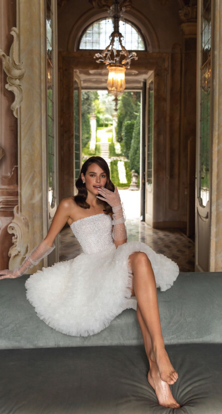 In questa foto una modella indossa un abito da sposa Pollardi 2023 corto, con microbalze. Seduta su una panca, indossa anche dei guanti in tulle 