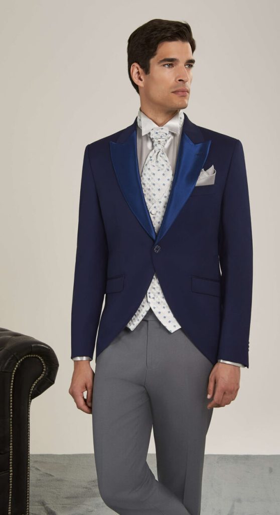 In questa foto un modello indossa un tight Andrea Versali 2023 composto da: giacca di colore bluette con revers a lancia in satin a contrasto; gilet e plastron di colore bianco con motivo a rombi; pochette di colore grigio perla; pantaloni di colore grigio antracite