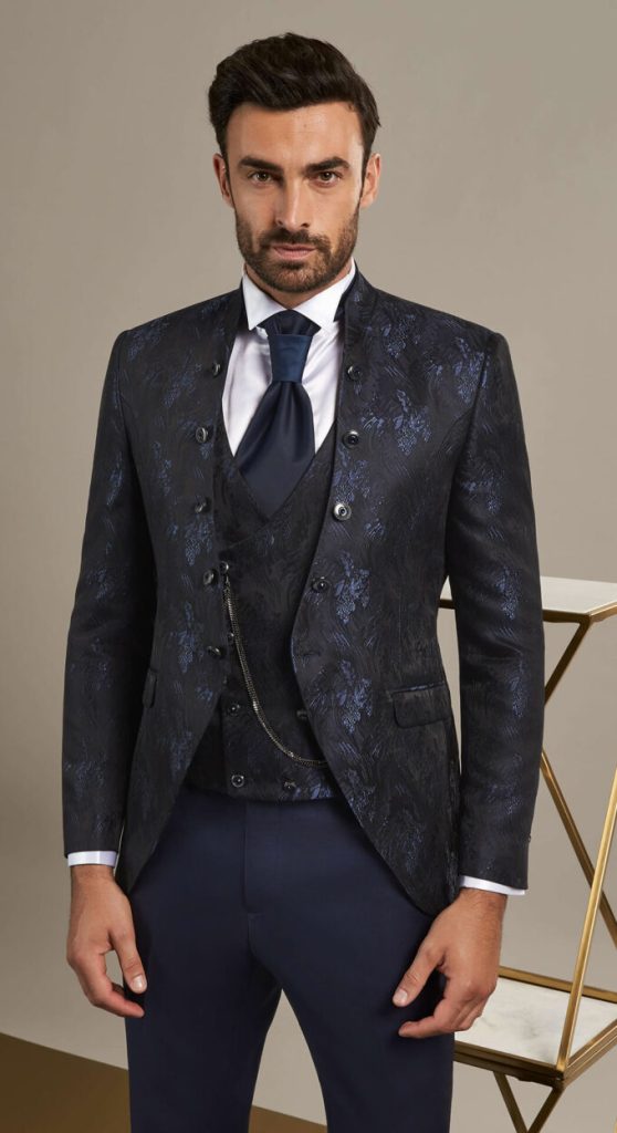 In questa foto un modello indossa un abito da cerimonia uomo con giacca coreana e gilet doppiopetto in tessuto damascato di colore bluette e nero; cravatta in satin di colore blu scurso; pantaloni abbinati