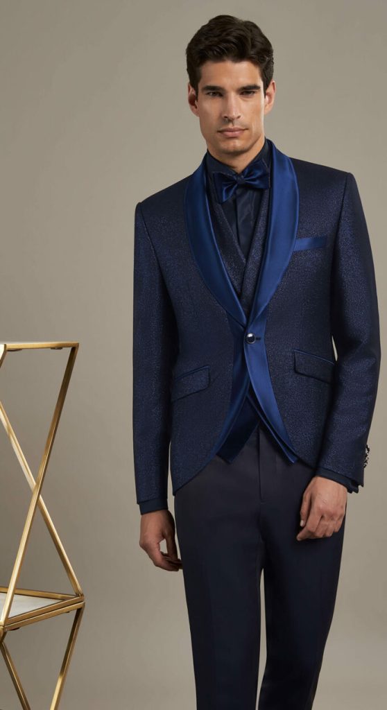 In questa foto un modello indossa uno smoking Andrea Versali 2023 con giacca di colore blu con motivo glitter tono su tono, revers in satin a contrasto, gilet abbinato, papillon in satin di colore blu scuro