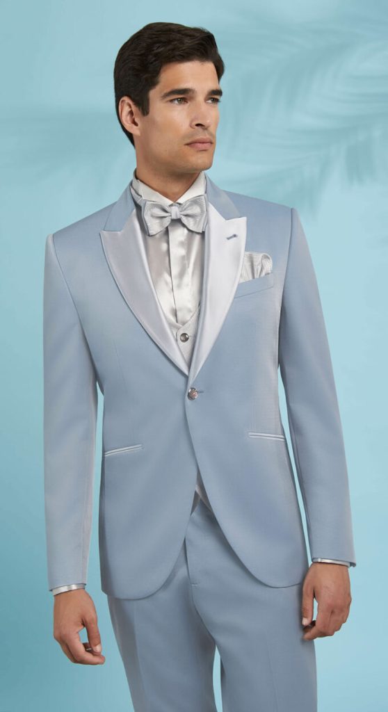 In questa foto un modello indossa un abito da cerimonia uomo di colore azzurro cielo e con giacca dai revers a lancia di colore grigio perla abbinati alla camicia, al gilet, al papillon e alla pochette nel taschino