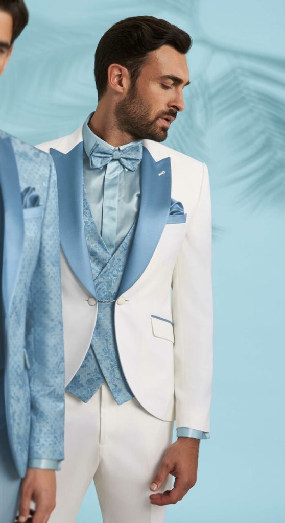 In questa foto un modello indossa uno smoking con giacca bianca e revers a lancia di colore azzurro cielo abbinati al gilet damascato, alla camicia, al papillon e alla pochette