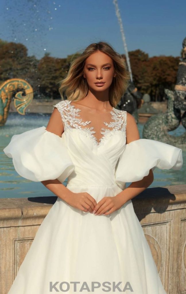 In questa foto una modella fotografata davanti ad una fontana a Parigi e a mezzo busto indossa un abito da sposa Iryna Kotapska ad A in chiffon con corpetto drappeggiato, maniche balloon e bretelle in pizzo ramage