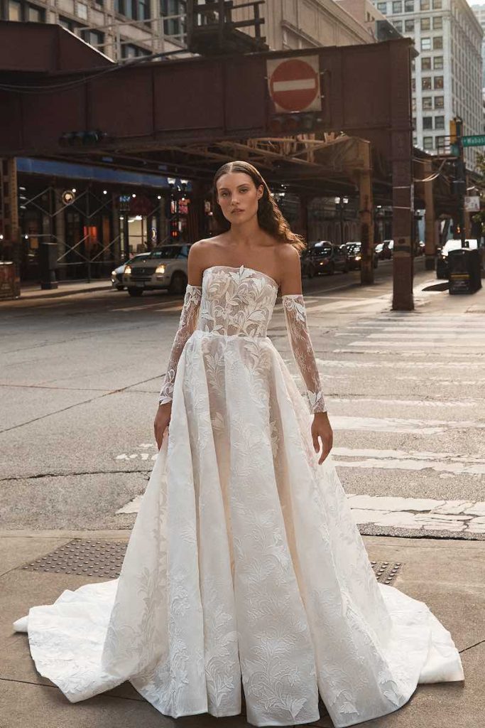 In questa foto una modella indossa un abito da sposa La Premiere Bridal 2023 dalla linea ad A in taffetà damascato. Il bustier steccato e trasparente con scollatura dritta riprende lo stesso ricamo presente anche sulle maniche in tulle rimovibili