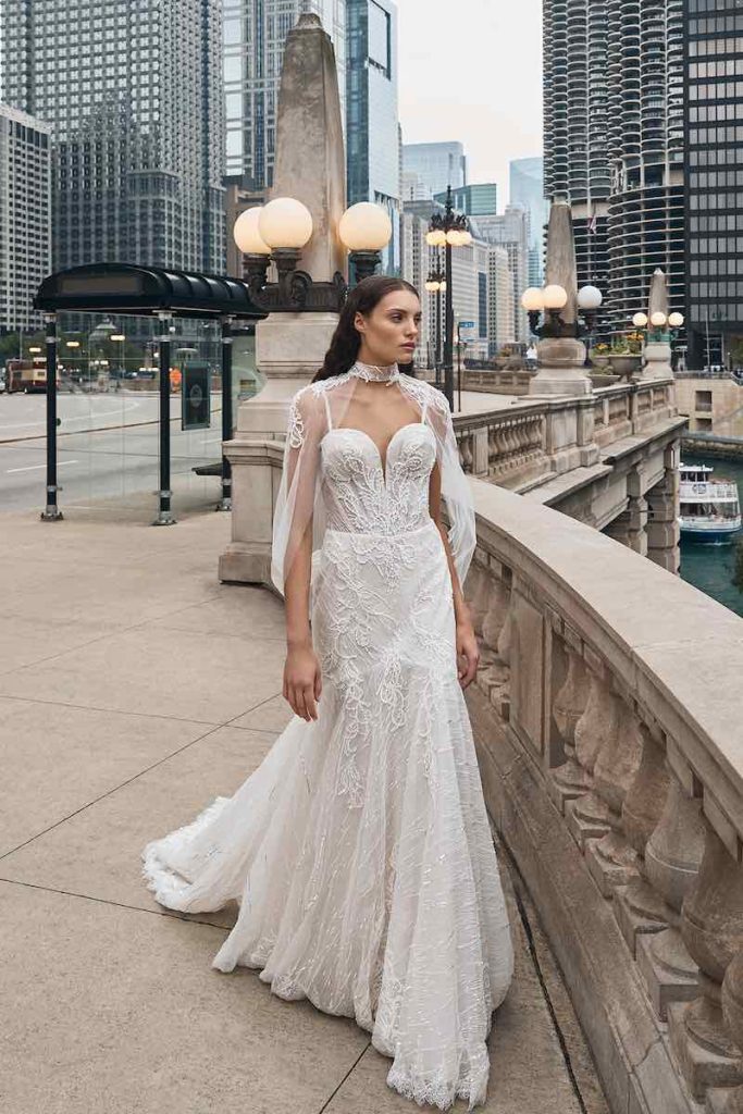 In questa foto una modella indossa un abito da sposa La Premiere Bridal 2023 a mezza sirena in pizzo con ricami di perline, bustino steccato, scollo a cuore con bretelline. Il look è completato da una cappa in tulle ricamato fermato da un collare gioiello e che scende fino alle spalle