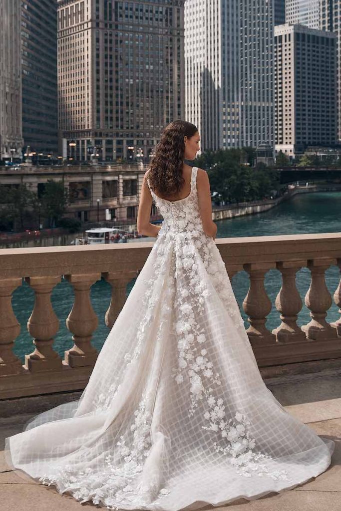 In questa foto una modella ritratta di spalle indossa un abito da sposa La Premiere Bridal 2023 ampio con coda con uno strato di tessuto stampato a scacchi e, sopra, uno strato di tulle con applicazioni di fiori 3D