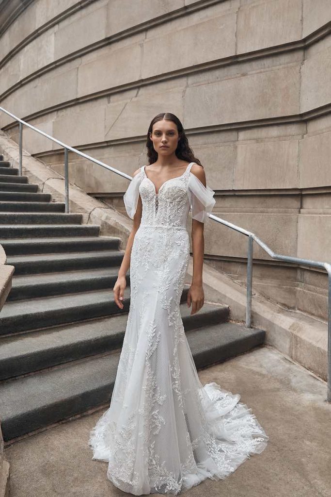 In questa foto una modella indossa un abito da sposa La Premiere Bridal 2023 a mezza sirena in tulle ricamato con un leggero scollo a V e maniche corte in tulle che scendono morbide sulle spalle