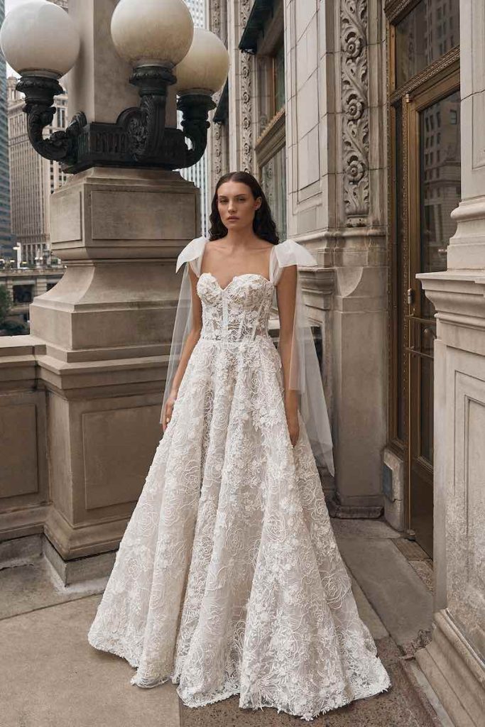 In questa foto una modella indossa un abito da sposa La Premiere Bridal 2023 dalla linea ad A interamente in pizzo 3D, bustier steccato con scollatura a cuore e fiocchi di tulle che scendono sulle braccia