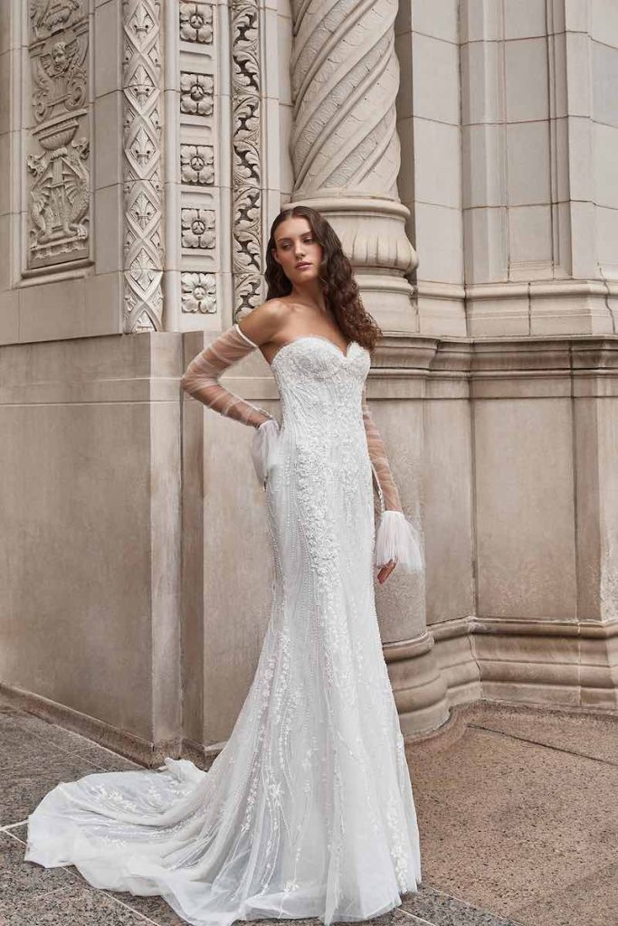 In questa foto una modella indossa un abito da sposa La Premiere Bridal 2023 a mezza sirena in tulle e organza con ricami di perline, scollo a cuore e maniche rimovibili a campana con dettagli