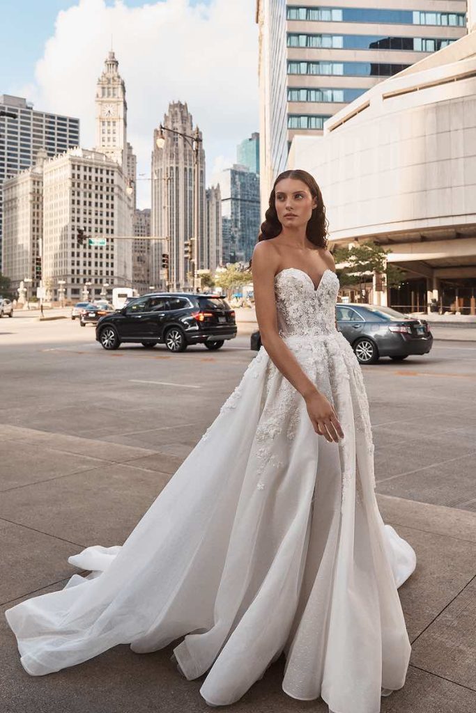 In questa foto una modella indossa un abito da sposa La Premiere Bridal 2023 ampio in organza con scollo a cuore e bustino decorato con fiori 3D che scendono ramage sulle lunghezze