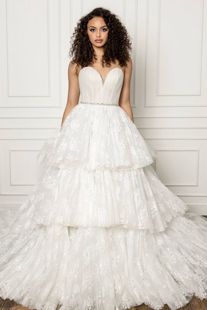 In questa foto una modella indossa un abito da sposa ampio in pizzo a balze, scollatura a cuore e cinturino gioiello stretto in vita