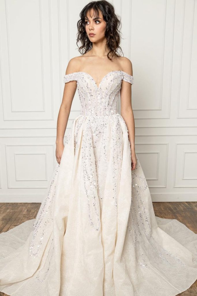 In questa foto una modella indossa un abito da sposa La Premiere Bridal 2023 dalla linea ad A colore avorio con scollatura a barchetta e ramage di paillettes colore argento 