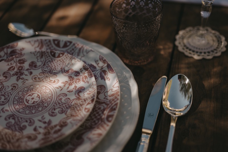 In questa foto il dettaglio di una mise en place di matrimonio con tavolo in legno nudo, piatti con decorazione di maioliche siciliane di colore rosso e posate d'argento