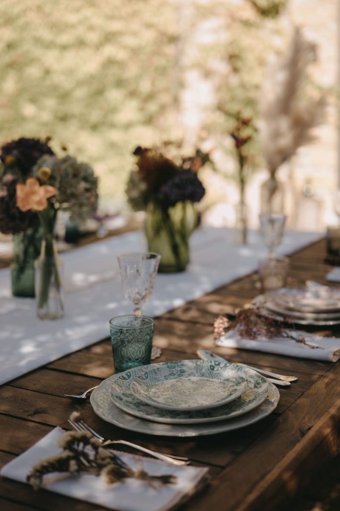 In questa foto il dettaglio di una mise en place di matrimonio con runner di lino di colore bianco, vasi di varie dimensioni con fiori di campo, piatti con decori di maioliche siciliane di colore verde e rosso e bicchieri da acqua abbinati