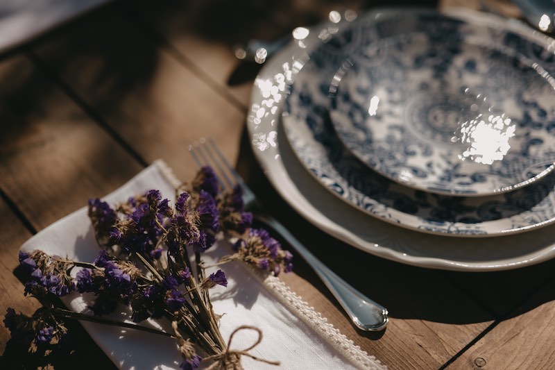 In questa foto il dettaglio di una mise en place di matrimonio con piatti con decori di maioliche siciliane di colore blu, tovagliolo in lino ricamato e fiori secchi di colore viola